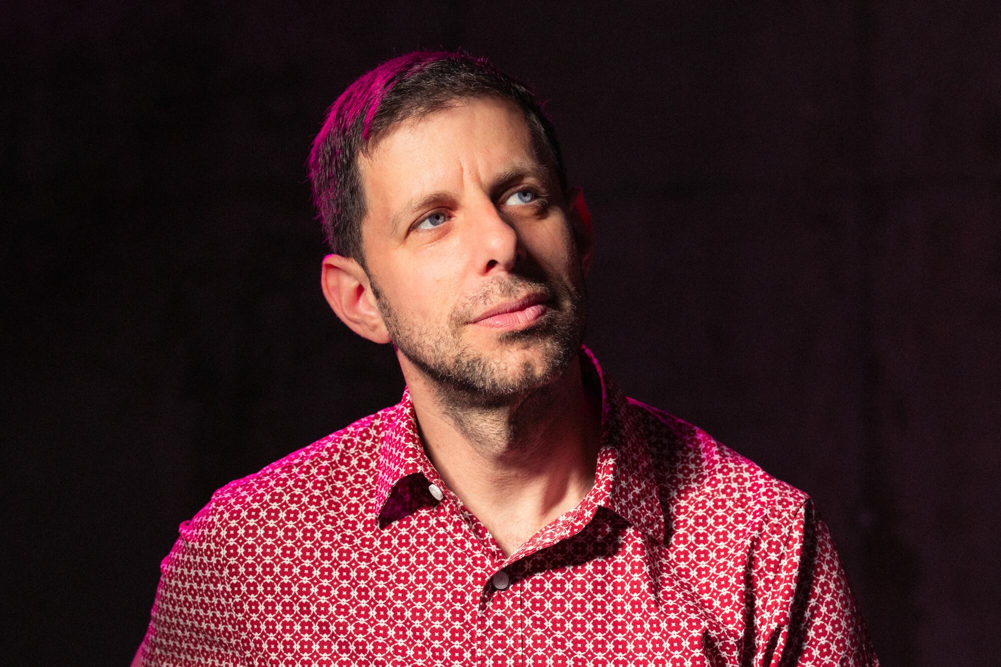 Yuval Sharon, in a red patterned shirt, sits in front of a black background and looks to the side.
