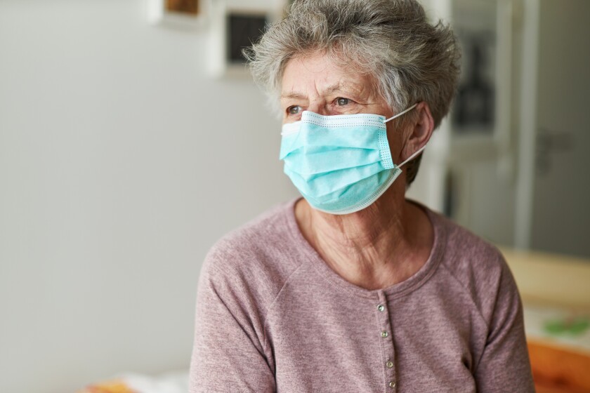 A senior citizen sits alone on her bed with a respirator or surgical mask and looks sadly and frightened out of the window and into the camera