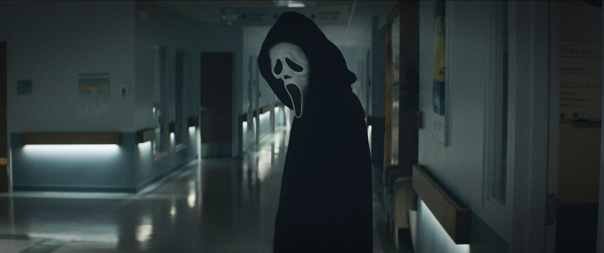 A person in a black cloak and white mask stands in a hallway