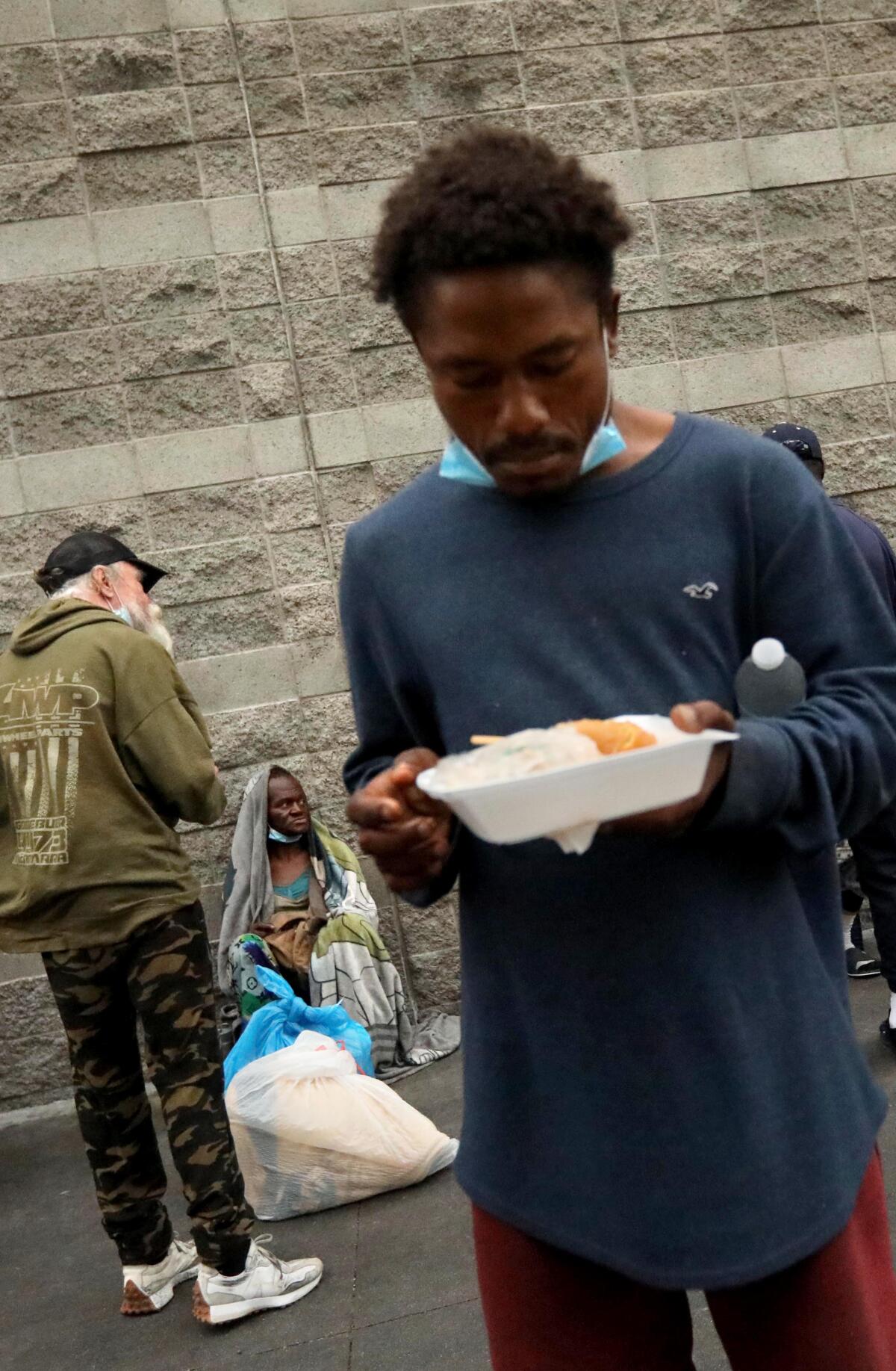 A homeless man prepares to eat his breakfast in a container while standing on the sidewalk.