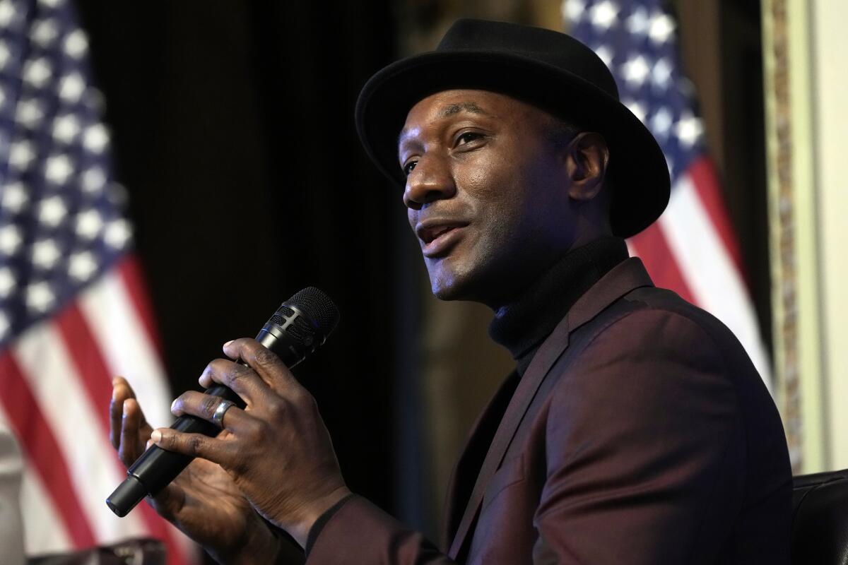 Aloe Blacc in a suit and a black hat speaks into a hand-held microphone in front of two American flags