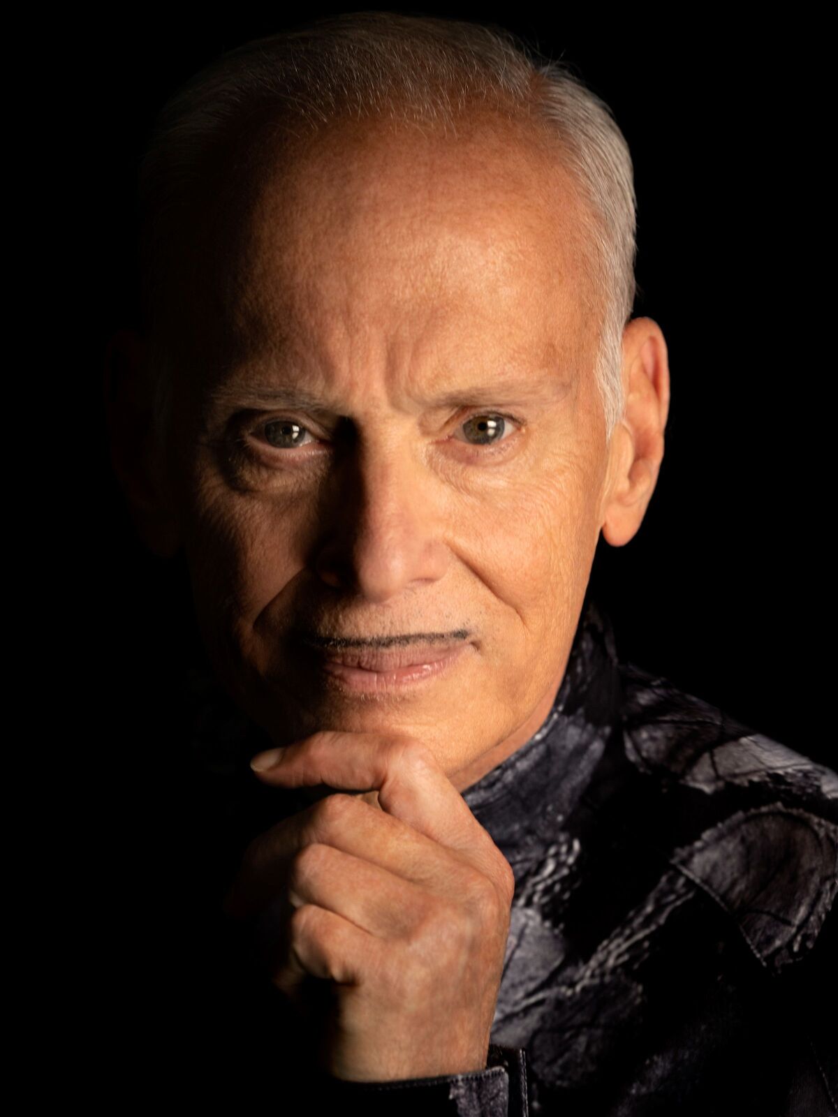 A man with gray hair and a pencil-thin mustache rests his chin on his hand