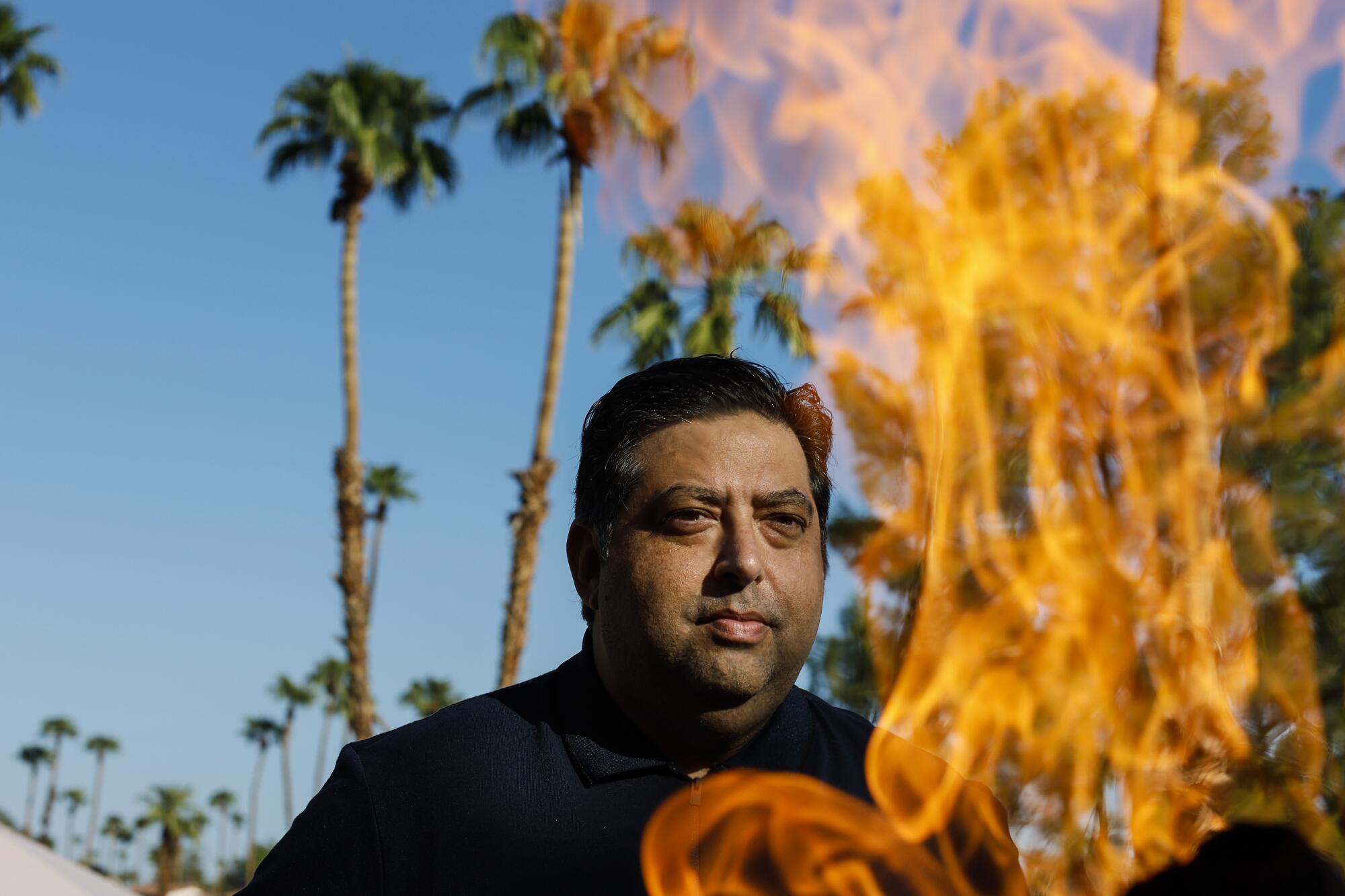 A man standing among palm trees beside a large daytime bonfire.
