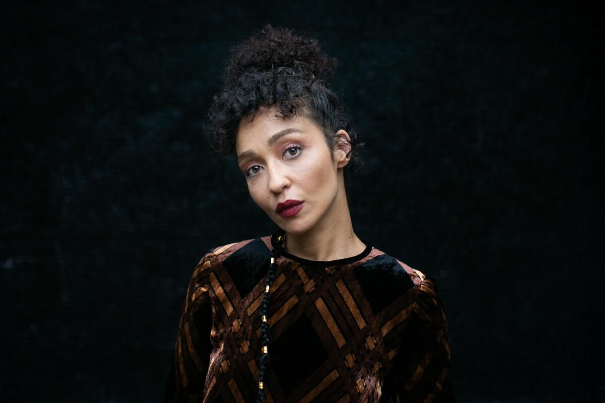 A portrait of Ruth Negga in a brown and gold striped dress against a dark background.