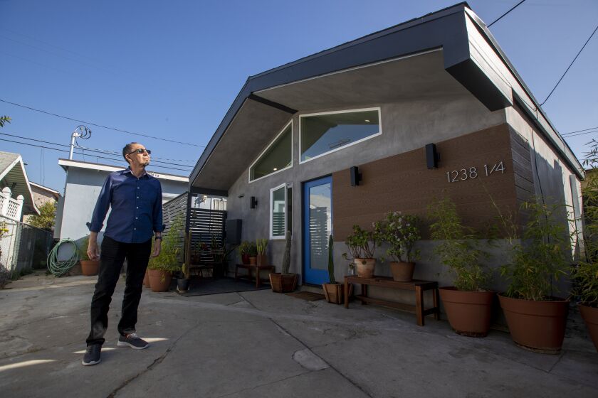 LOS ANGELES, CA - February 06: Landlord and architect Alexis Navarro stands in front of his "Casita L.A.," a new ADU he designed in East L.A. in the carport of an existing 3-unit property Saturday, Feb. 6, 2021 in Los Angeles. Navarro designed and built the 500-square-foot ADU over five months for $95,000. He designed it to be open and flexible, especially with the adjacent houses and no exterior views. The design appropriates the Mid-Century Post and Beam system of a high ceiling but using conventional wood-frame construction to lower the cost and ease of building. The open floor plan was meant to provide options for flexible uses with only the bathroom as a closed space. Moveable partitions, curtains or furniture for privacy could be used to divide into various combinations of uses according to the lifestyle, needs or size of family. An abundance of daylight thru skylights and opaque glass windows, adds to the openness of the small space. Low cost alternatives for plumbing, electrical and kitchen accessories helped to meet the budget goal of less than $100,000. Energy conservation strategies include high ceilings with openable skylights to create an airflow to release the hot air to the outside. Large glazing clerestory windows face towards the north orientation and on the south exposure there are no openings to reduce the heat load. The raw concrete floor adds to the temperature stability of the house in the summer. Ariel Gomez-Hernandez and John Velasco and their dog, Lando, are renting the unit for $1,650 right now, and they are both working from home. (Allen J. Schaben / Los Angeles Times)