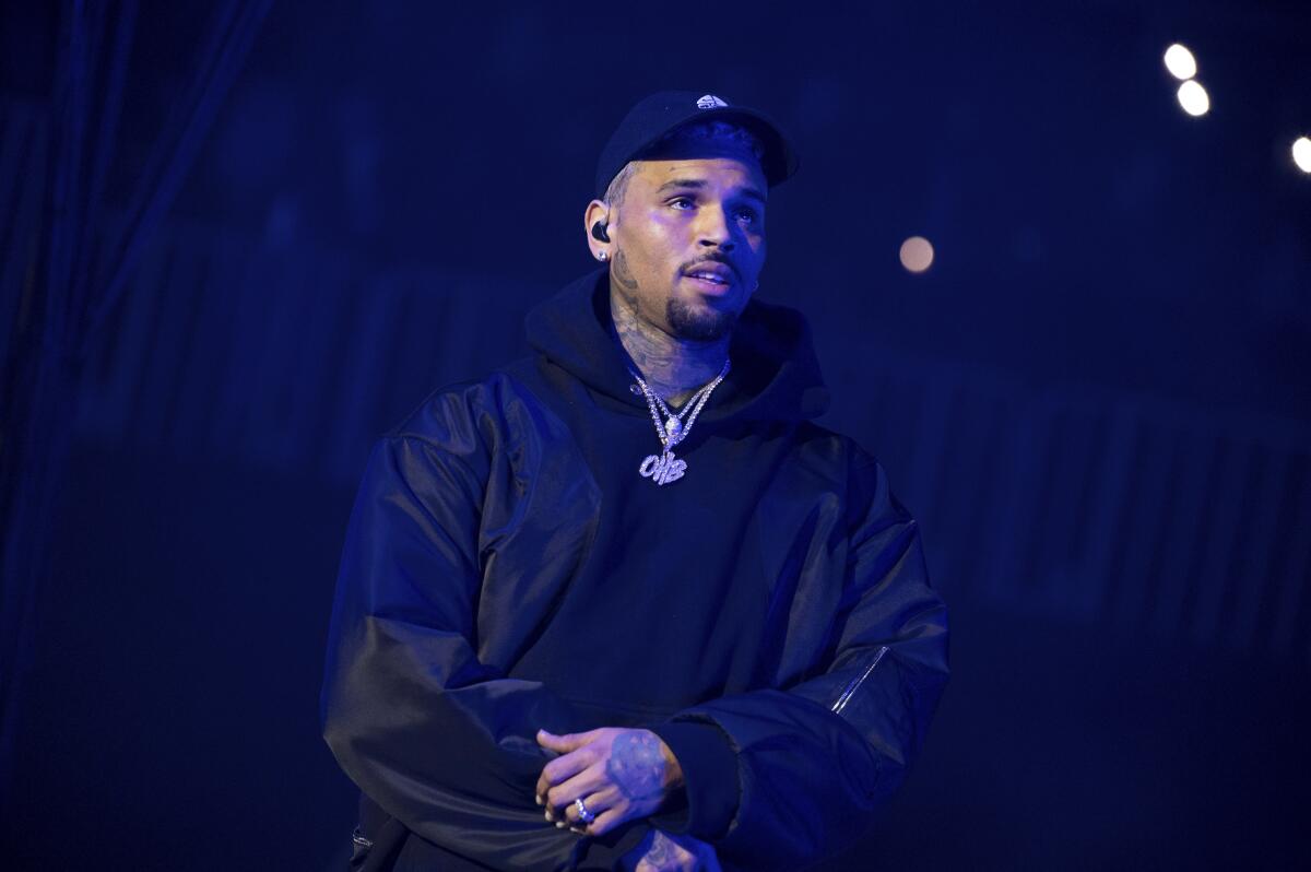 Chris Brown wears a black cap, hoodie and leather jacket while standing in front of a black backdrop.