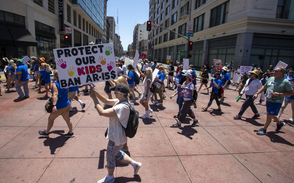 A crowd walking through an intersection, one with a sign reading "Protect our kids/Ban AR-15$," with a dollar sign for the S.