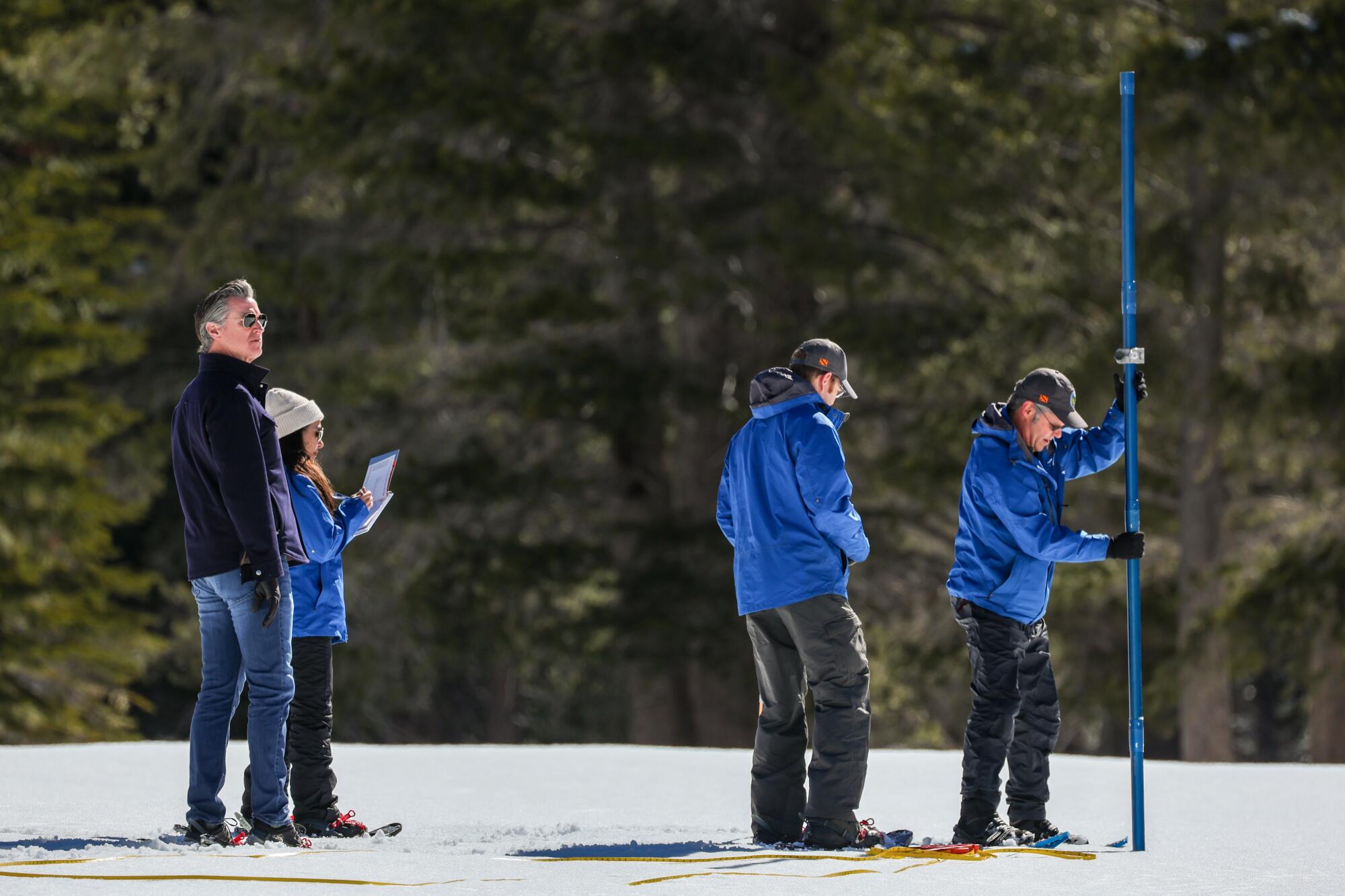 Gavin Newsom in the mountains with three people in blue jackets as one sticks a blue pole into the snow