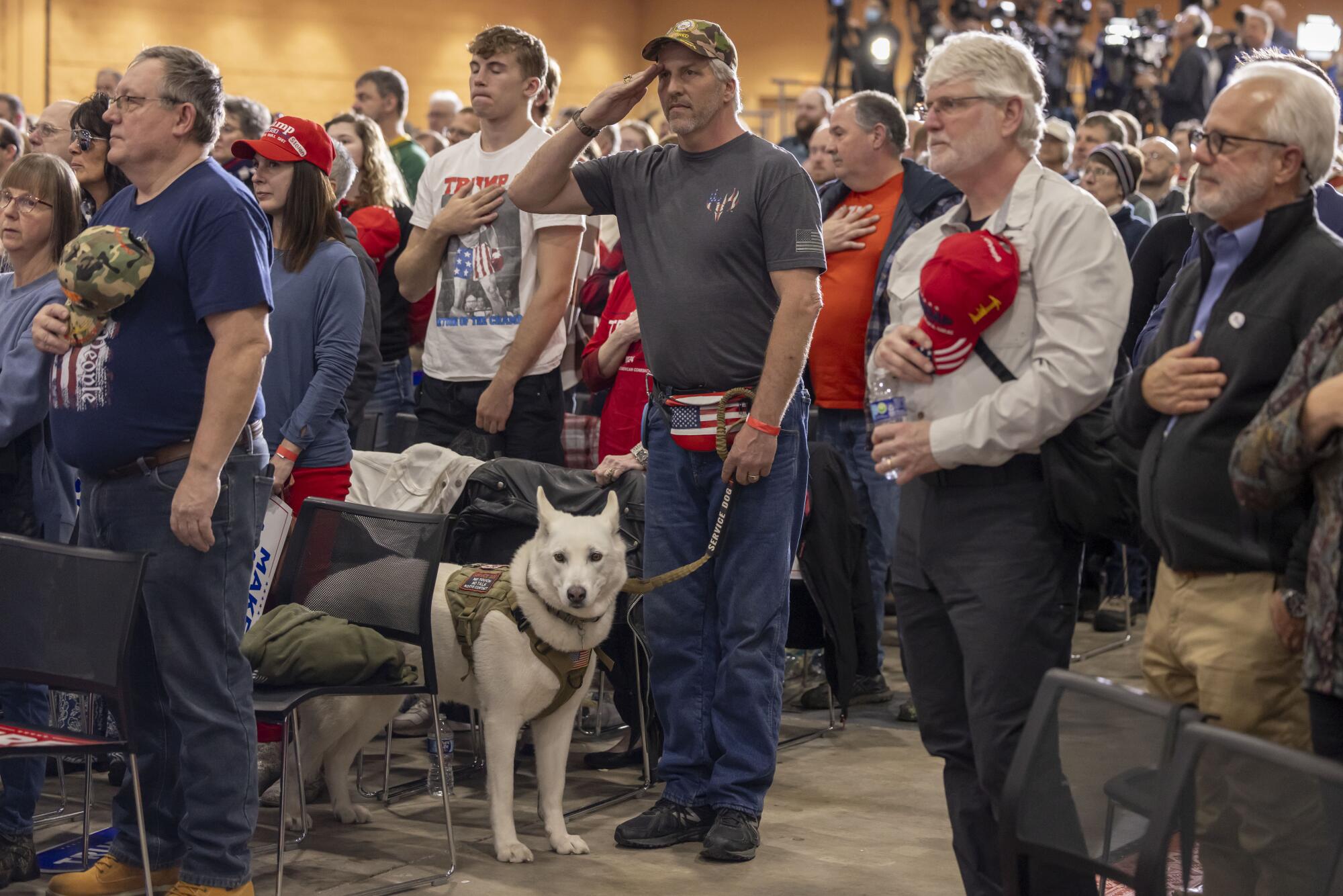 Uma multidão em pé, alguns com chapéus ou camisas Trump, a maioria com as mãos e chapéus no coração, enquanto um homem com um cachorro branco saúda