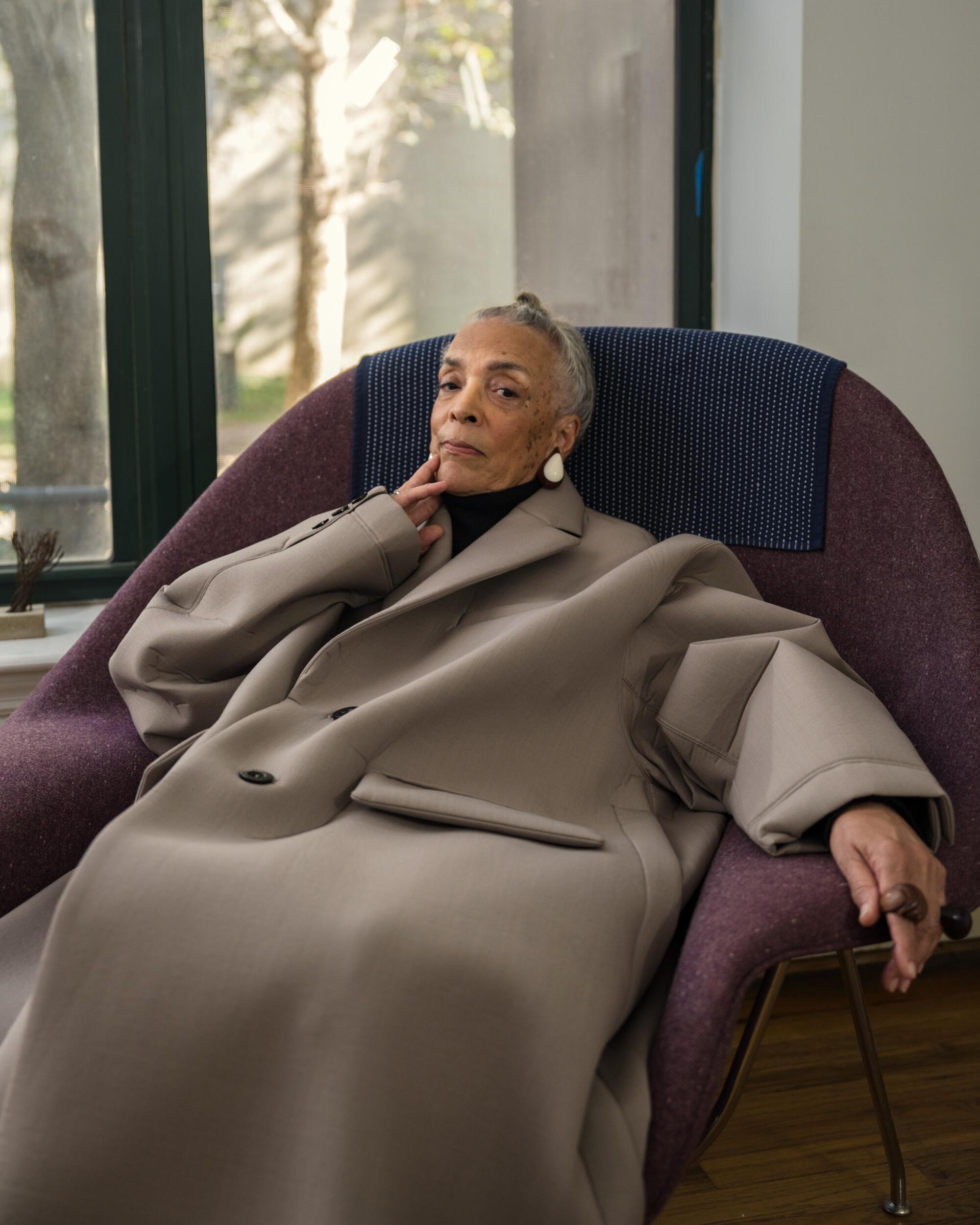 A woman, wearing an oversized brown jacket, sits on a purple chaise.