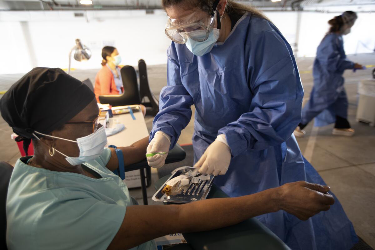 A doctor in protective gear picks up instruments to use for drawing blood from a seated patient