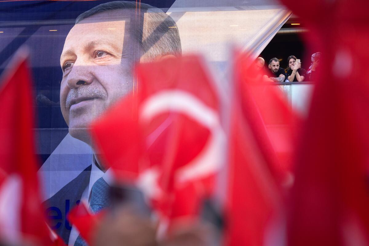 Framed by red flags, people watch from a cafe window at right, as a large Recep Tayyip Erdogan poster fills the left 