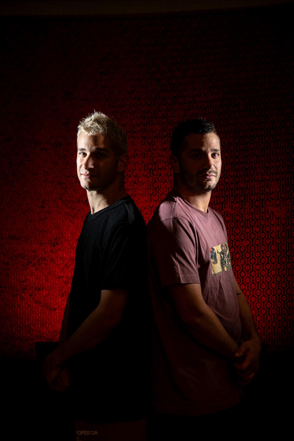Two men stand back to back in front of a deep red backdrop