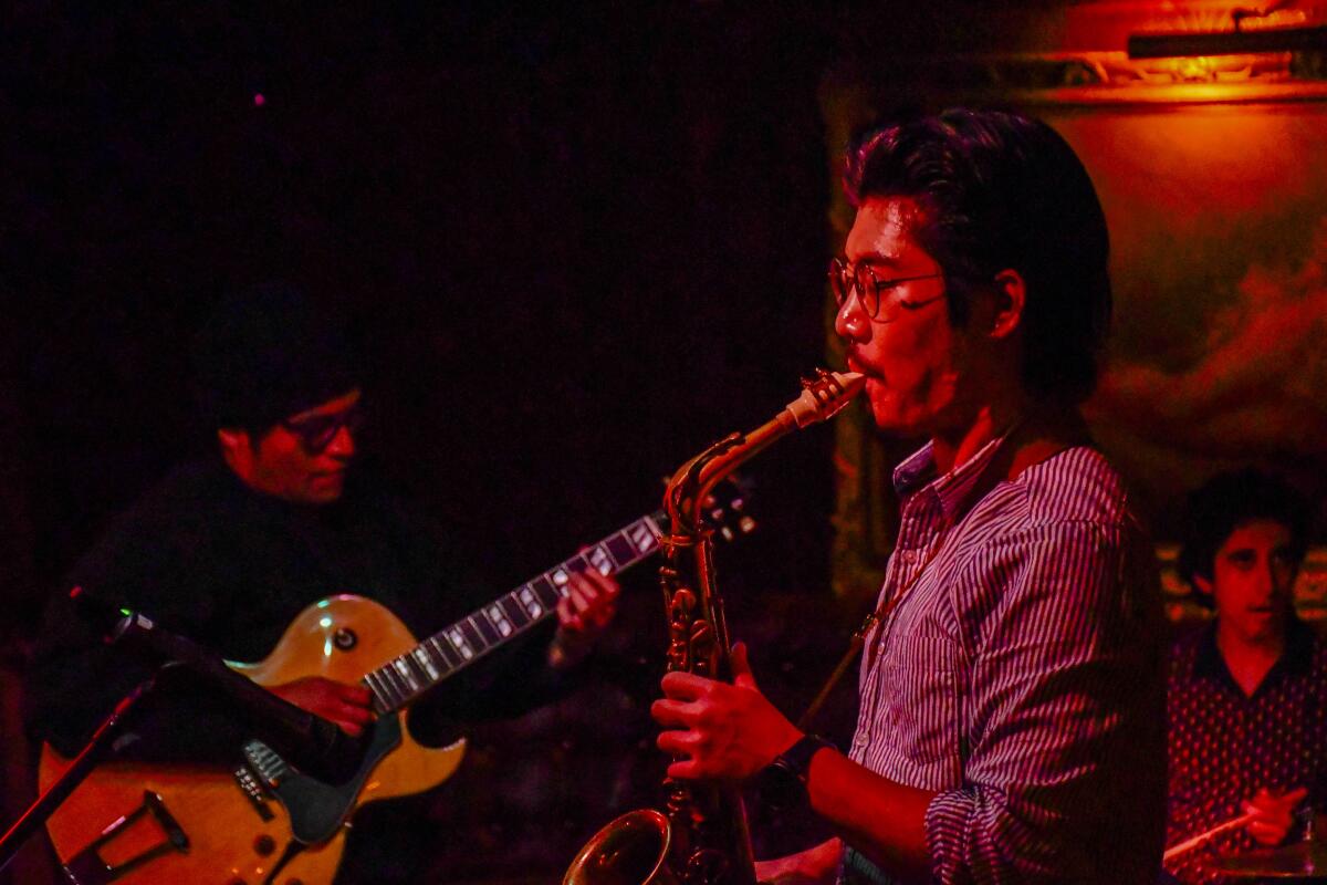 Musicians play guitar and saxophone in a red-lit room.