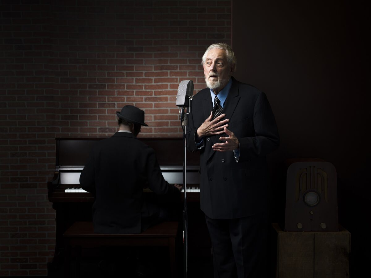 An elderly man in a suit speaks into an old-fashioned microphone onstage.