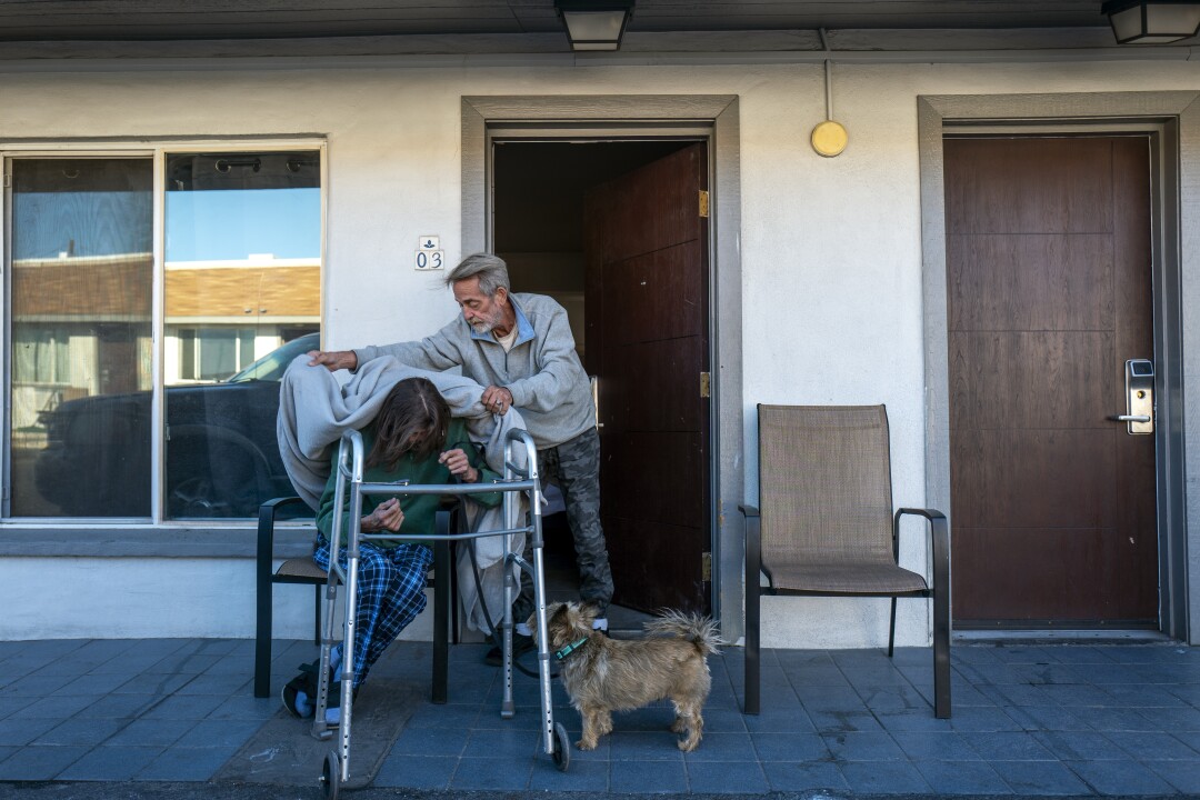 A man places a blanket over a seated man looking down, with a walker in front of him and a dog nearby.