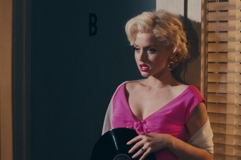 A woman with short, platinum blond hair wearing a pink dress, holding a vinyl record and leaning against a wall