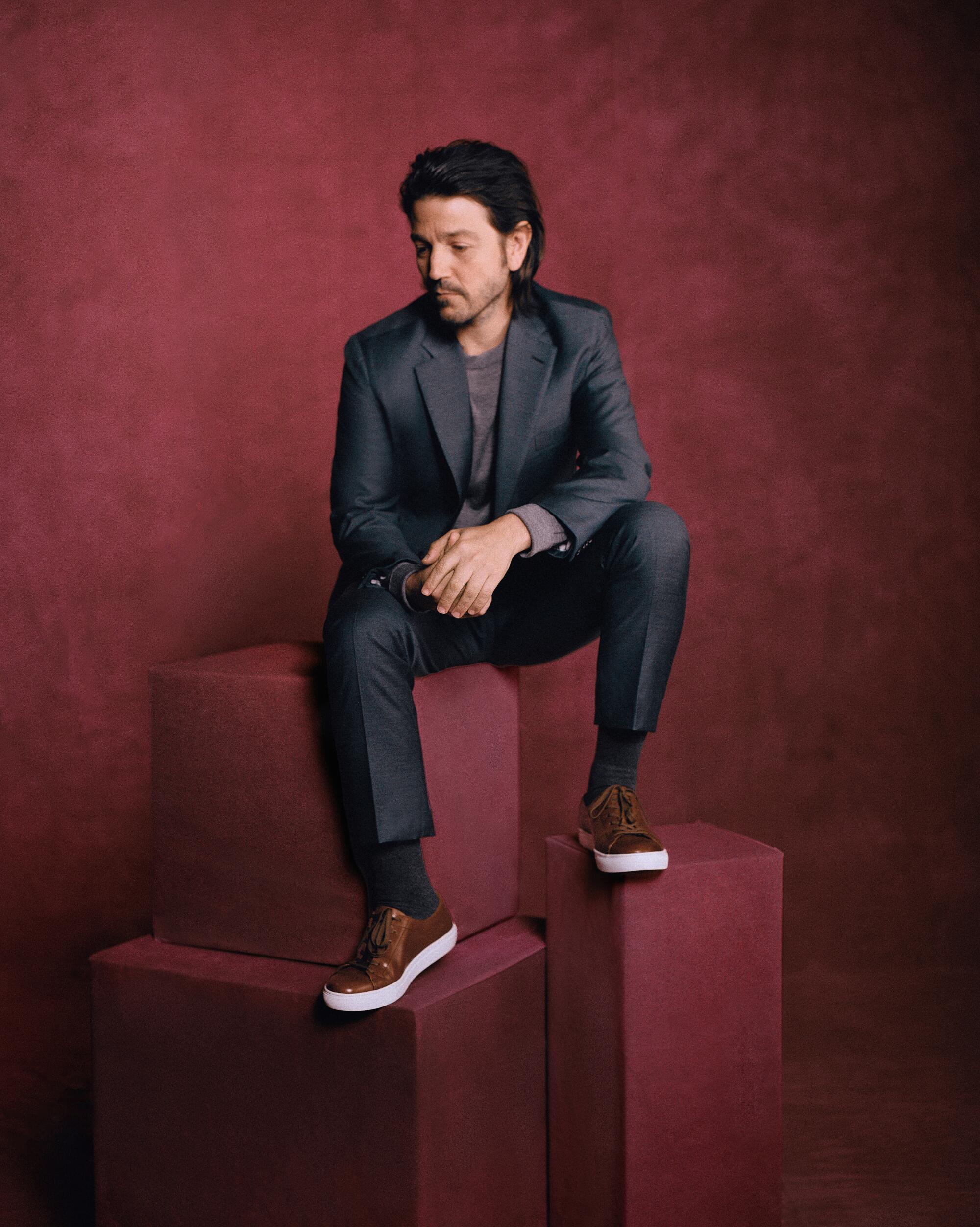Actor Diego Luna sits pensively on blocks before a burgundy background at L.A. Times HQ in 2023. 