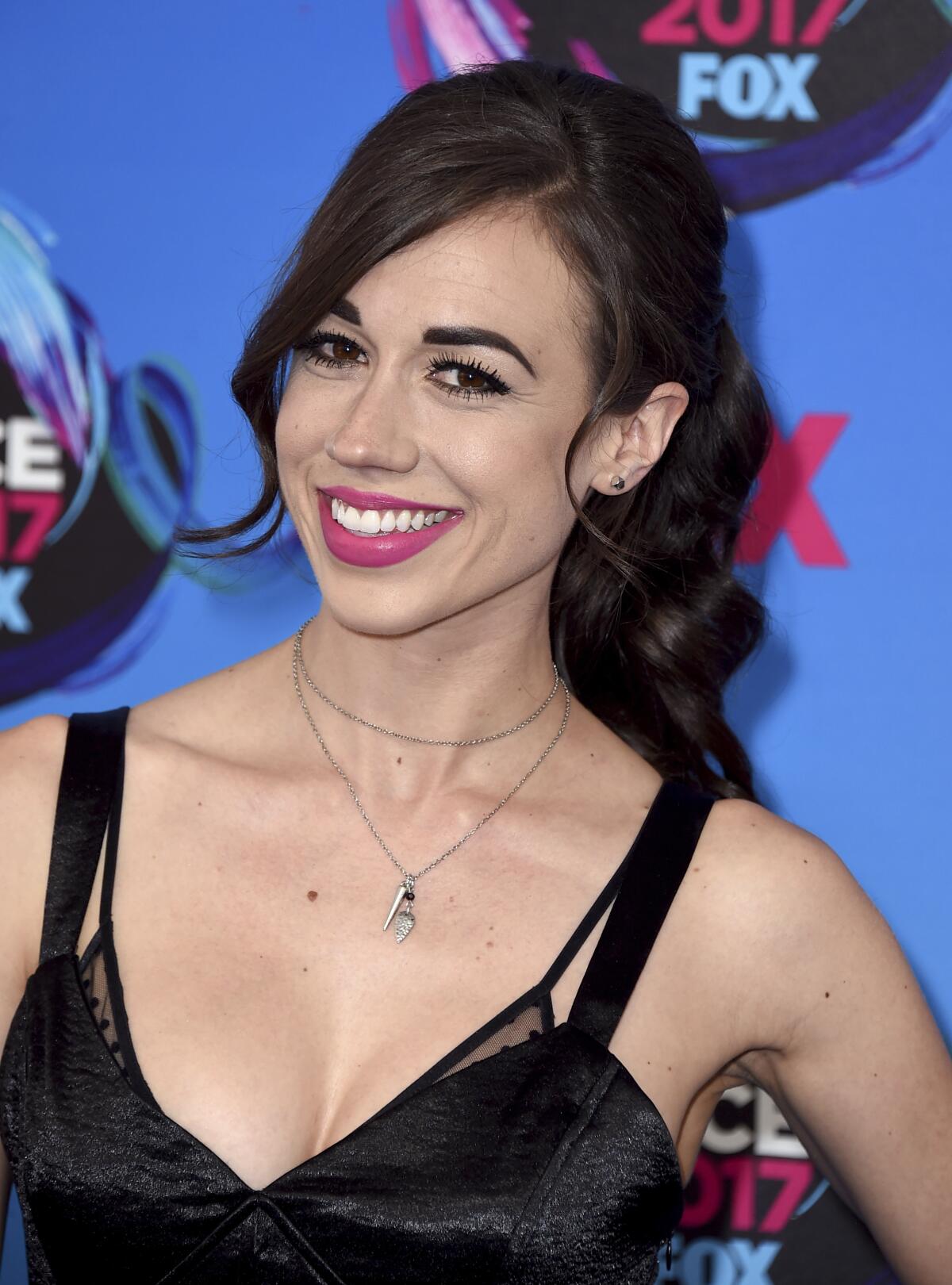 Colleen Ballinger with bangs and a ponytail wearing a strappy black dress against a blue backdrop