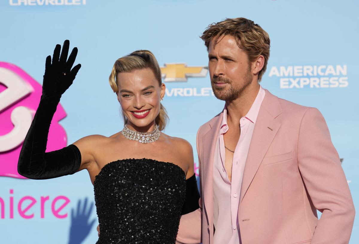 Margot Robbie is wearing a black dress and is smiling and waving next to Ryan Gosling, who is in a pink suit
