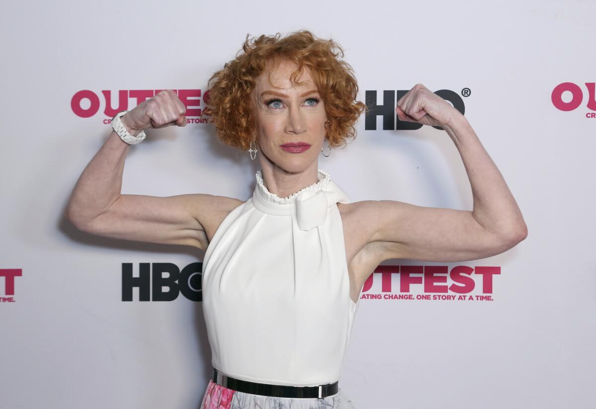 A woman in a sleeveless blouse flexes her arm muscles like a body builder and stares off into the distance