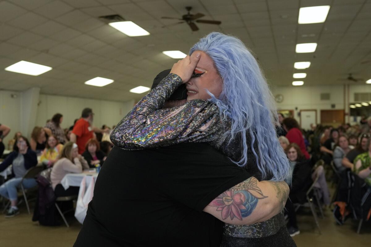 a drag queen with blue hair cries in front of an audience while embracing someone