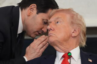 A man with dark hair, in a dark suit, leans down to whisper to a man with blond hair, in suit and red tie