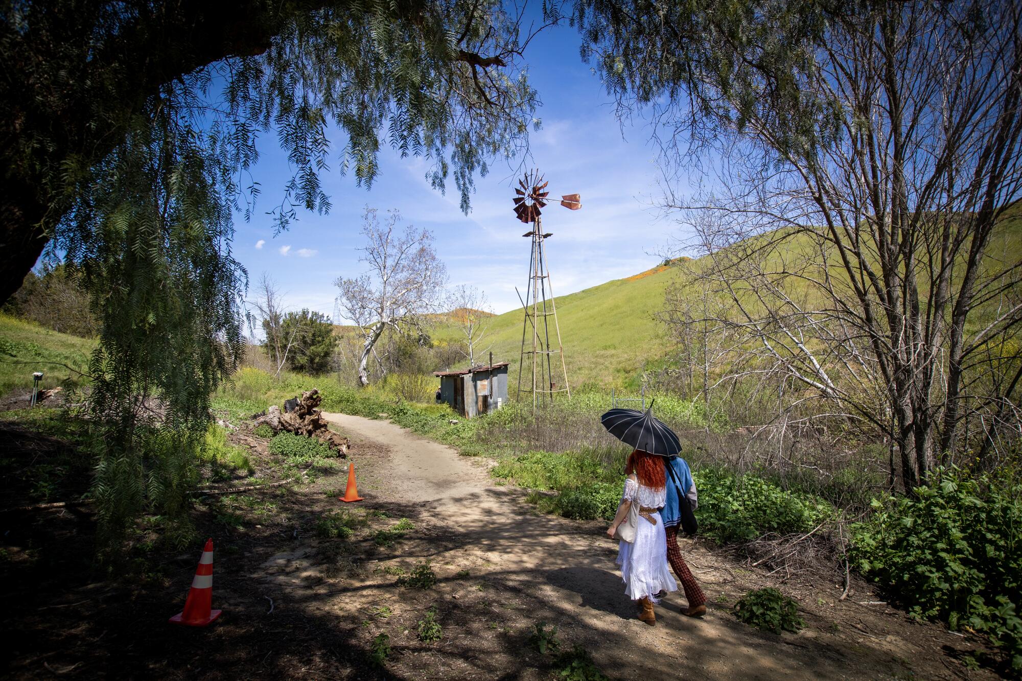 A couple huddled together under sun umbrella past a windmill on stilts; walking on a winding path between lush hills 