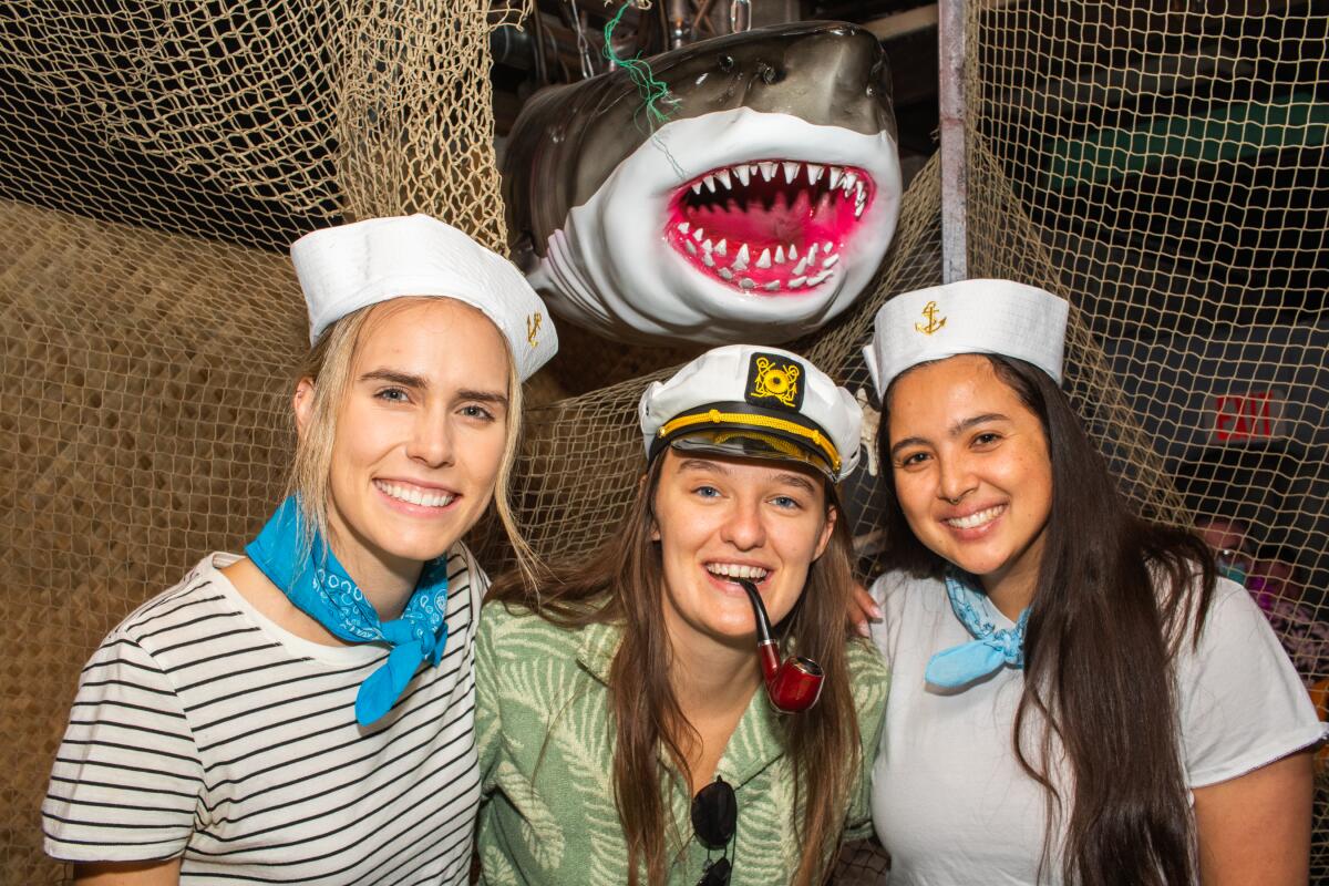 Two women in sailor tops and one wearing a captain's hat pose in front of a fiberglass shark.