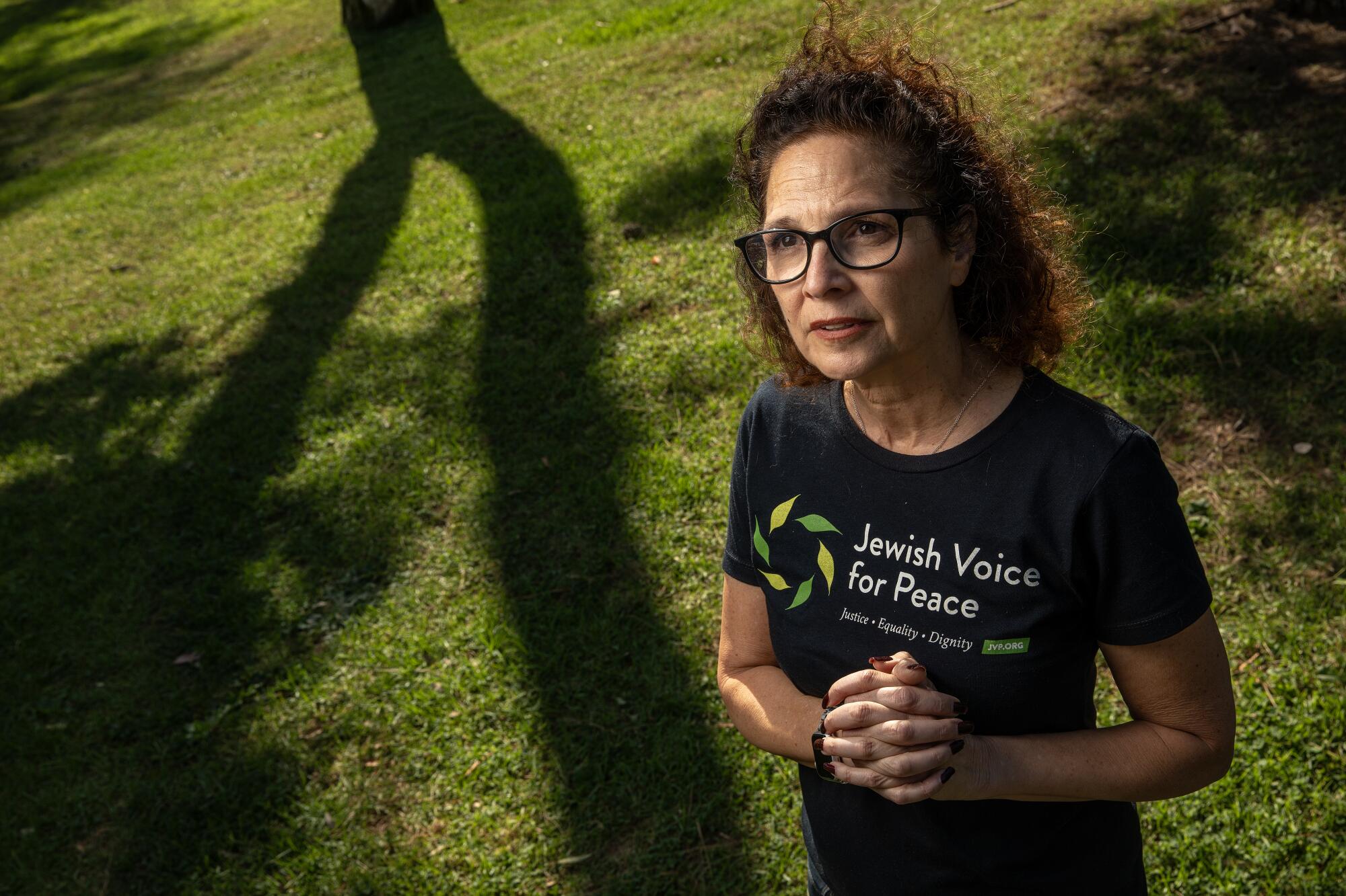 A woman in a black "Jewish Voice for Peace" T-shirt clasps her hands as she stands in grass, framed by the shadows of trees