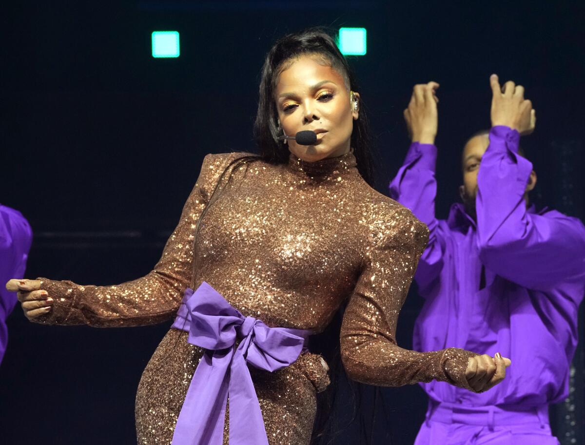 Janet Jackson onstage wearing a gold strong-shouldered jumpsuit with a purple bow at the waist