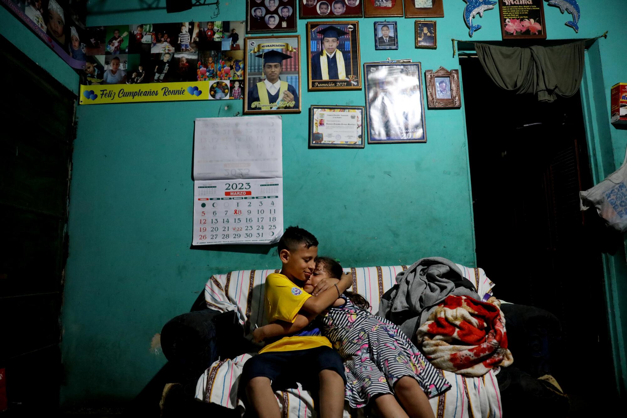 A boy and a girl embrace on a couch in front of a turquoise-colored wall adorned with pictures 