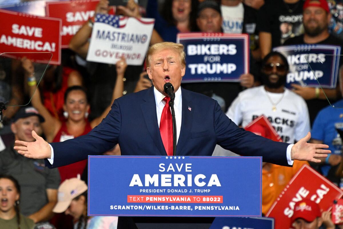 Ex-President Trump speaking at a lectern with a "Save America" sign as people in the crowd behind him hold various GOP signs.