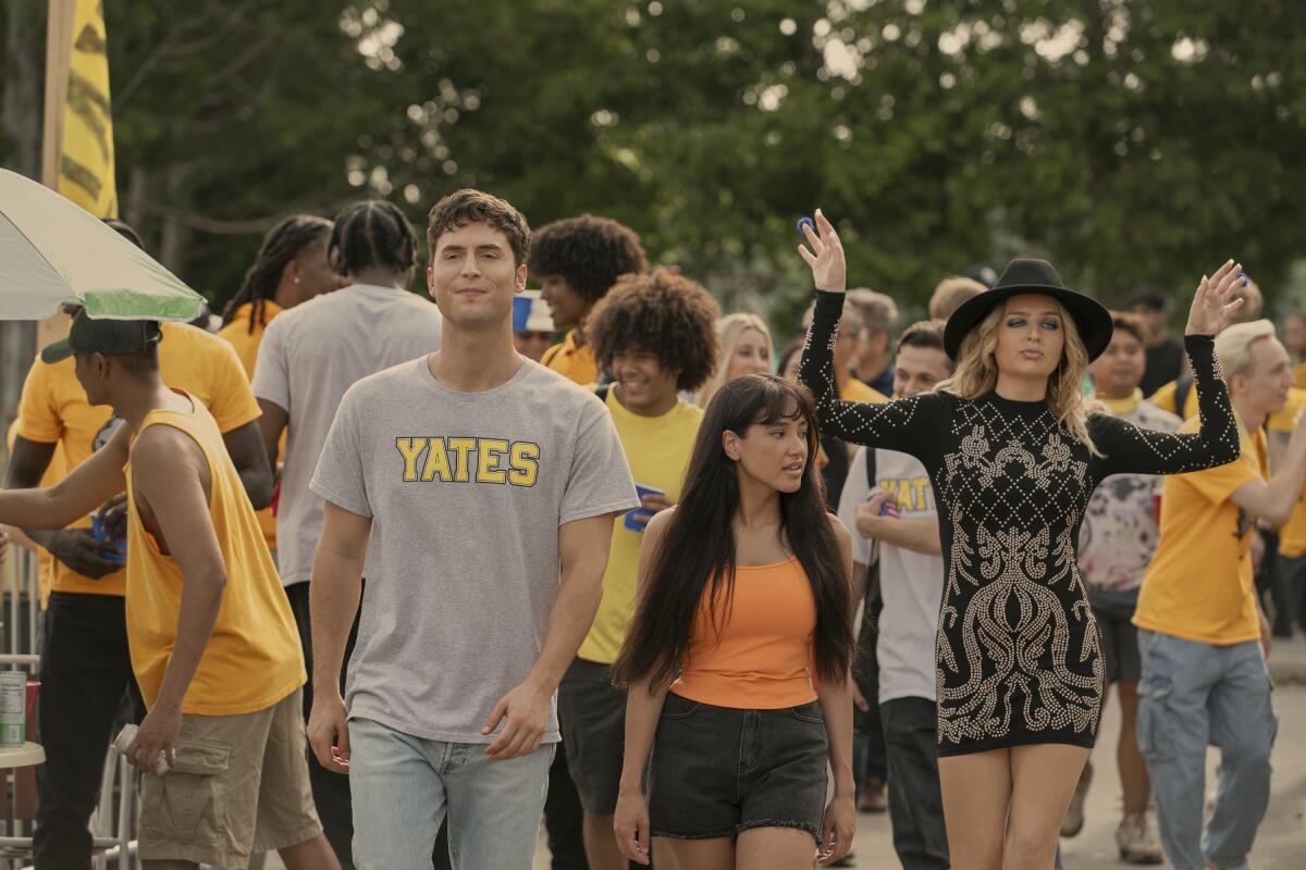 A man in a T-shirt and jeans walks with two women, one in an orange tank top and shorts, the other in a black dress and hat.