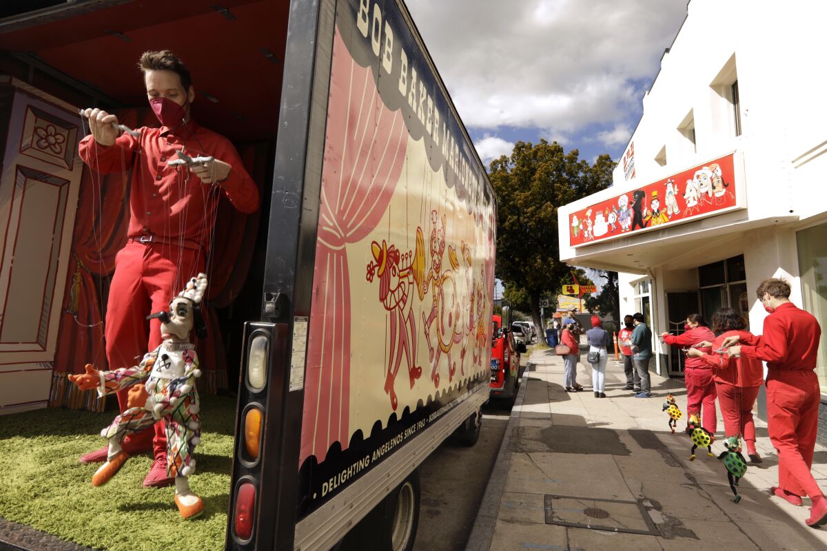 A man wearing red is seen manipulating a marionette of a dog on the back of a truck as other performers line a sidewalk.