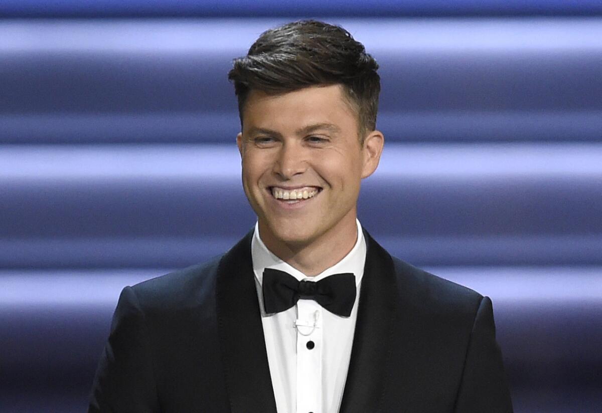 Colin Jost wearing a tuxedo and smiling against a blue background