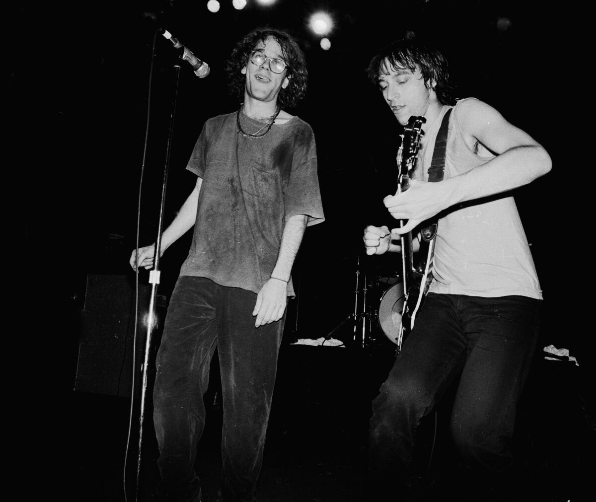 A black-and-white photo of two sweaty men, the one on the right holding a guitar, on stage