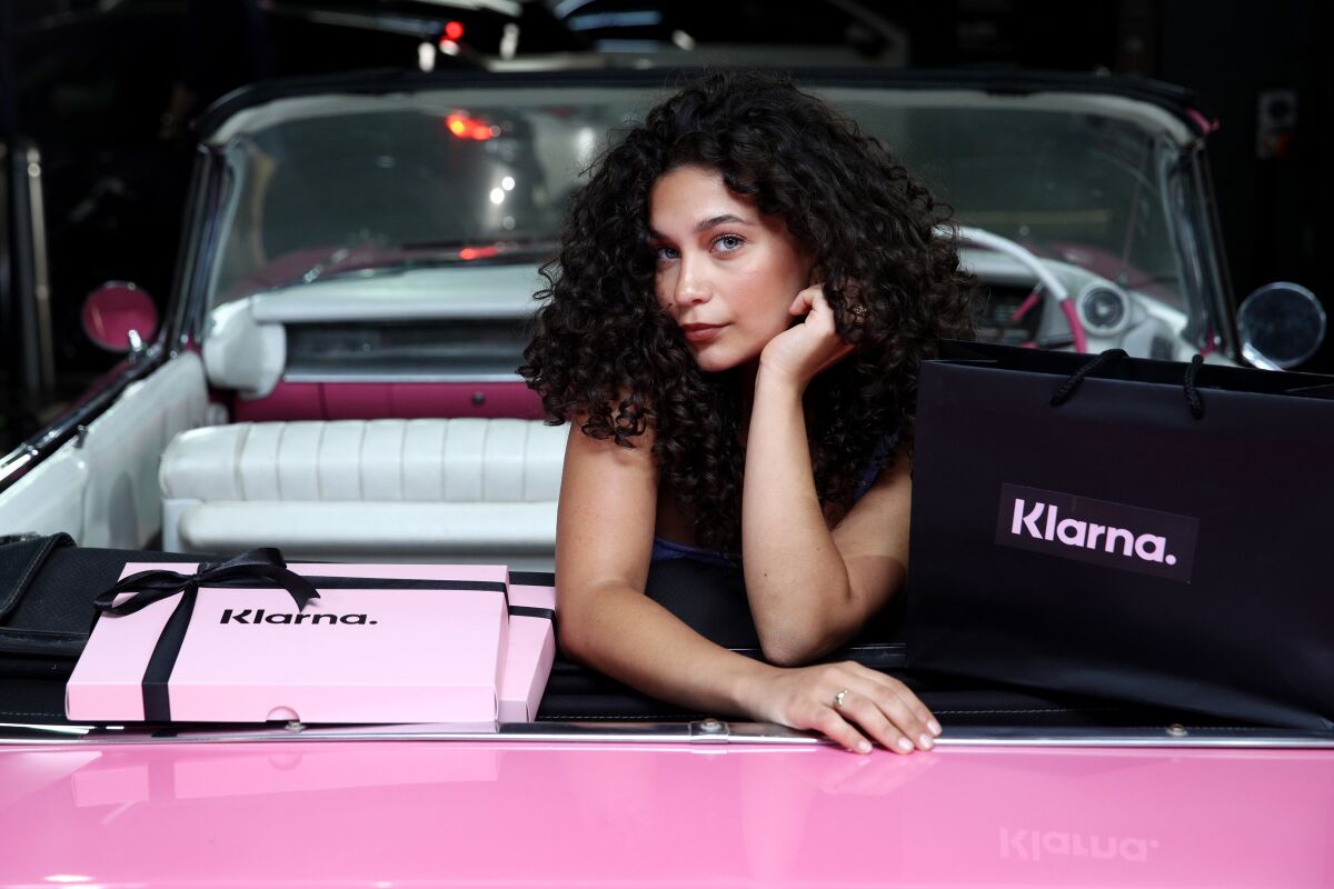 A woman with curly dark hair poses inside a pink Cadillac next to pink gift boxes and a black bag with the name Klarna