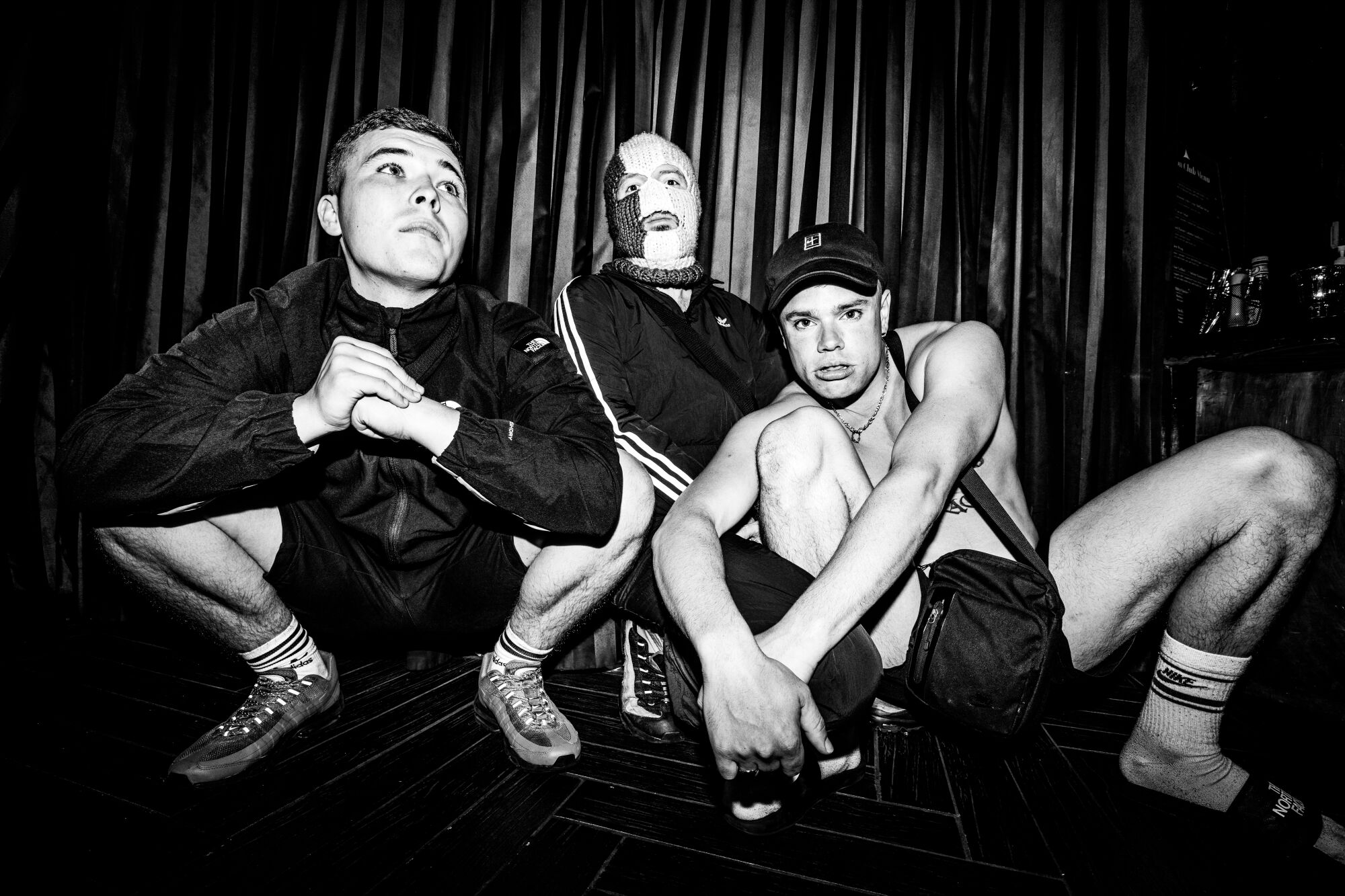 Three band members, one with a knit cap over his face, crouch down.