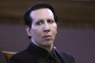 Marilyn Manson is sitting in a courtroom, looking to the left with a serious face while wearing a black suit 