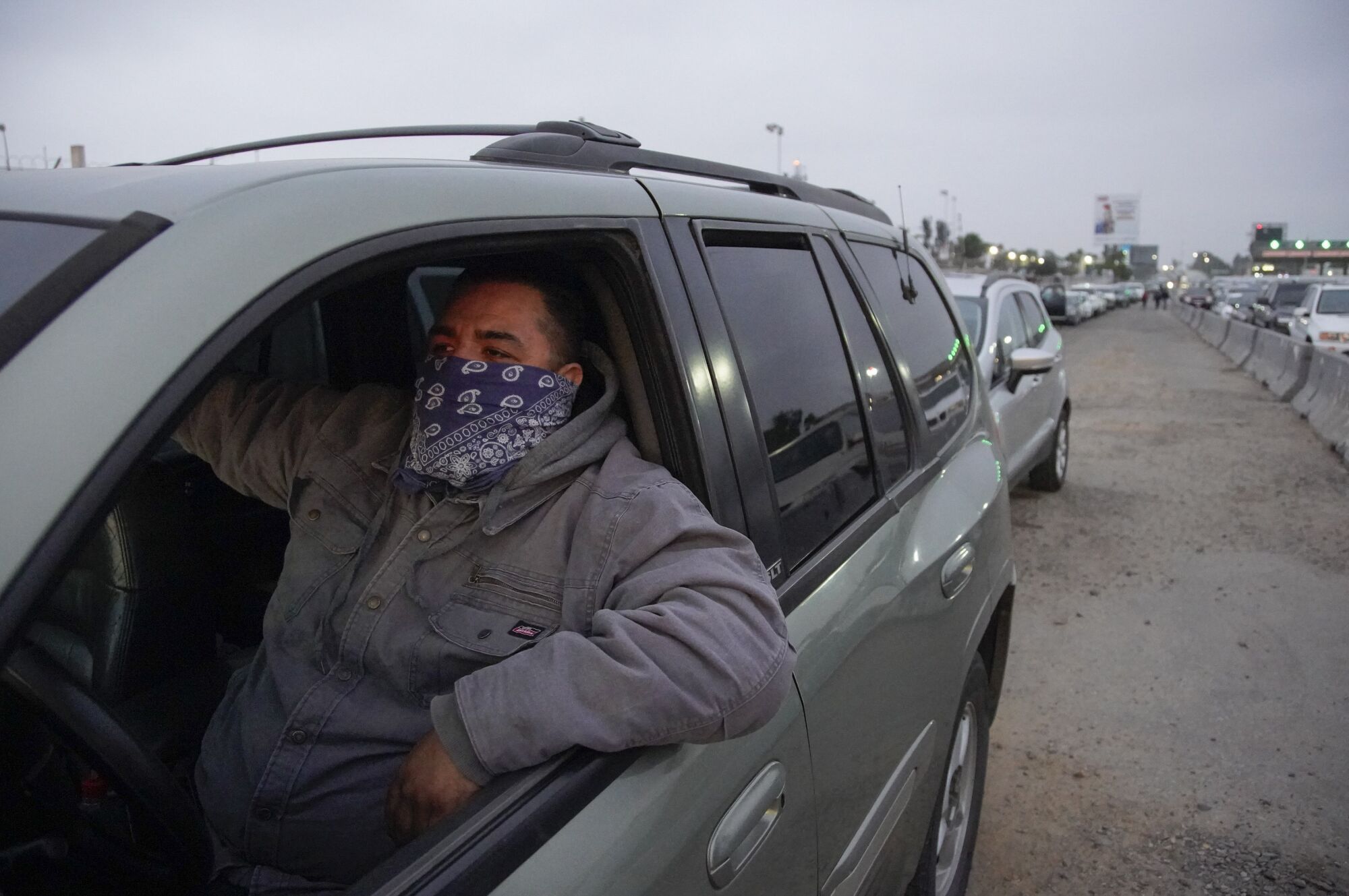 A man wearing a bandanna over his face waits in his car in a long line to cross the border