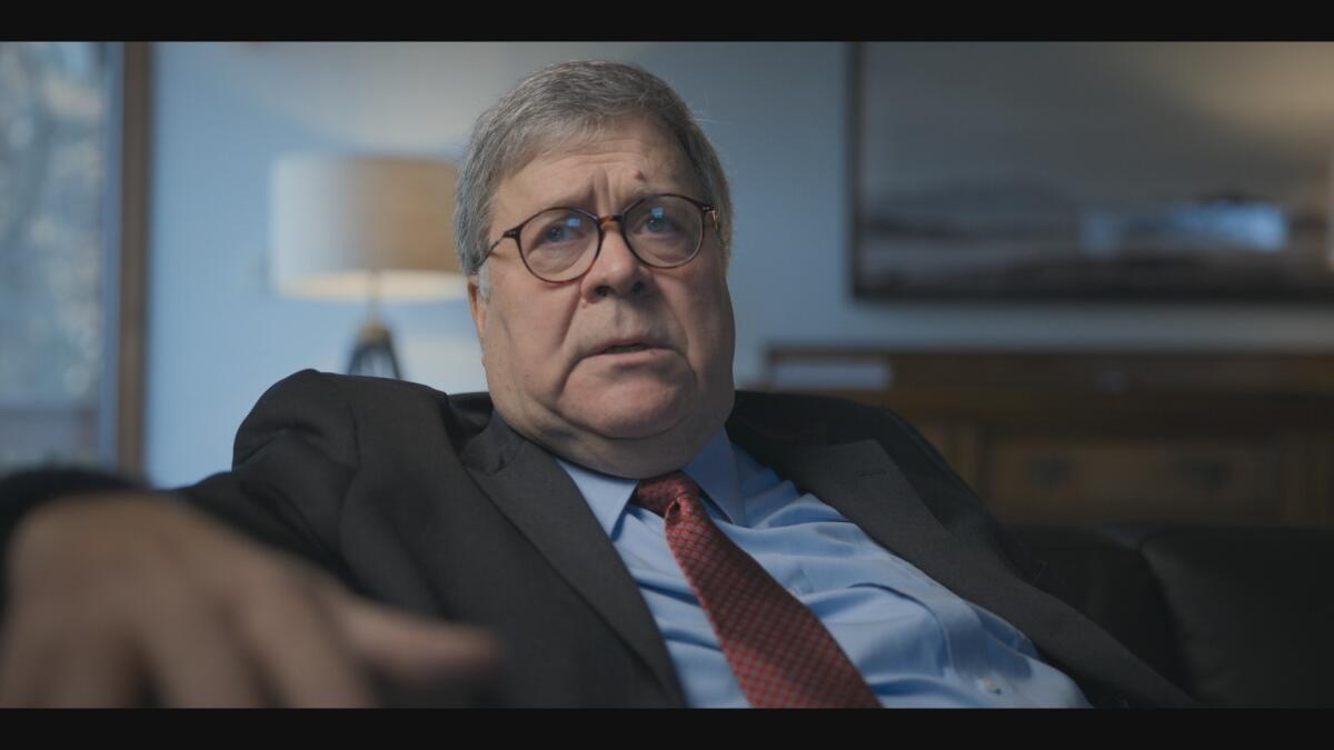 A man in brown-rimmed glasses, a blue shirt, orange tie and suit jacket sitting.