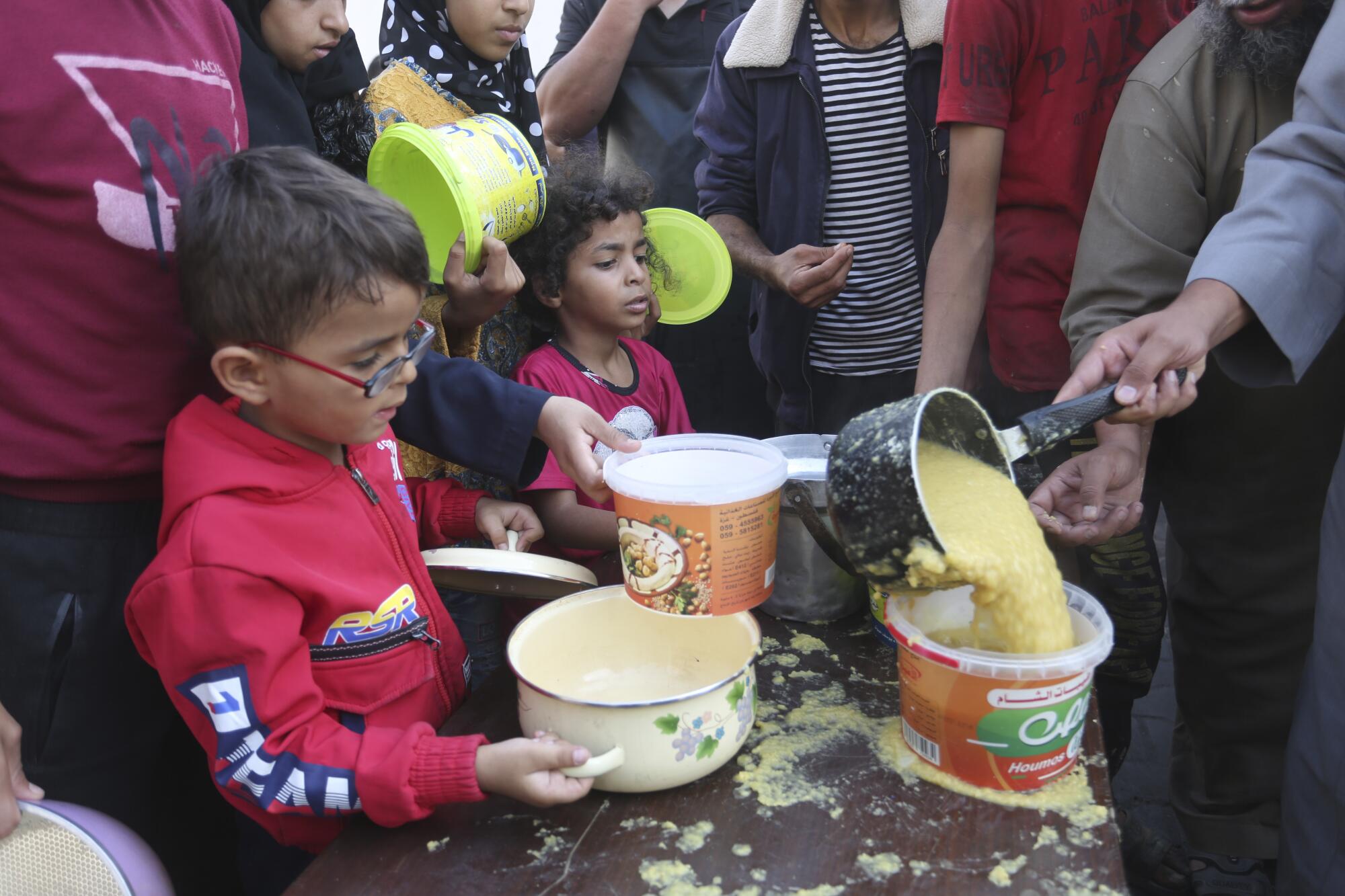 Children wait with adults as a person pours a yellow liquid into buckets 