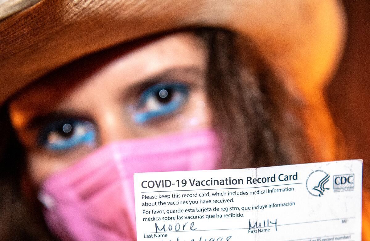 A woman in a cowboy hat and mask holds up a COVID vaccination card