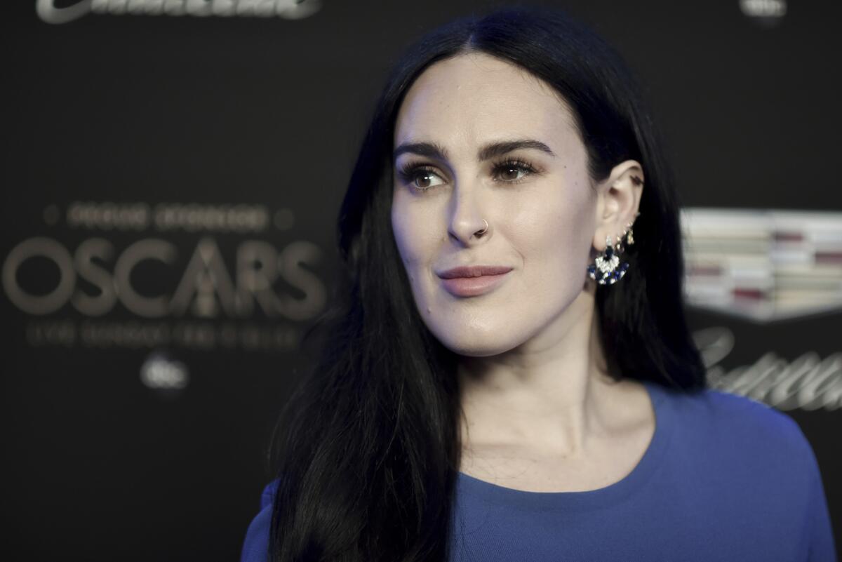 Rumer Willis wears a blue dress as she poses for photos in front of an Oscars backdrop