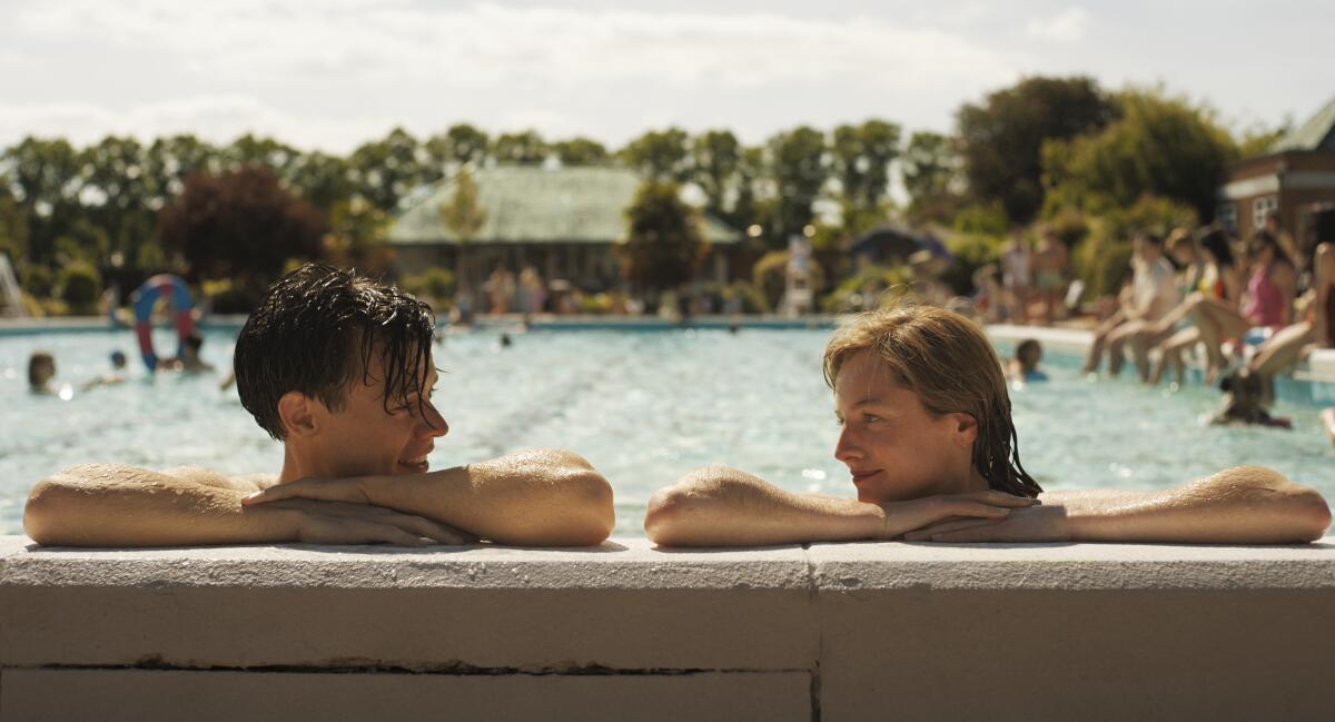A man and a woman in a swimming pool, resting their arms on the edge of the pool, look at each other.