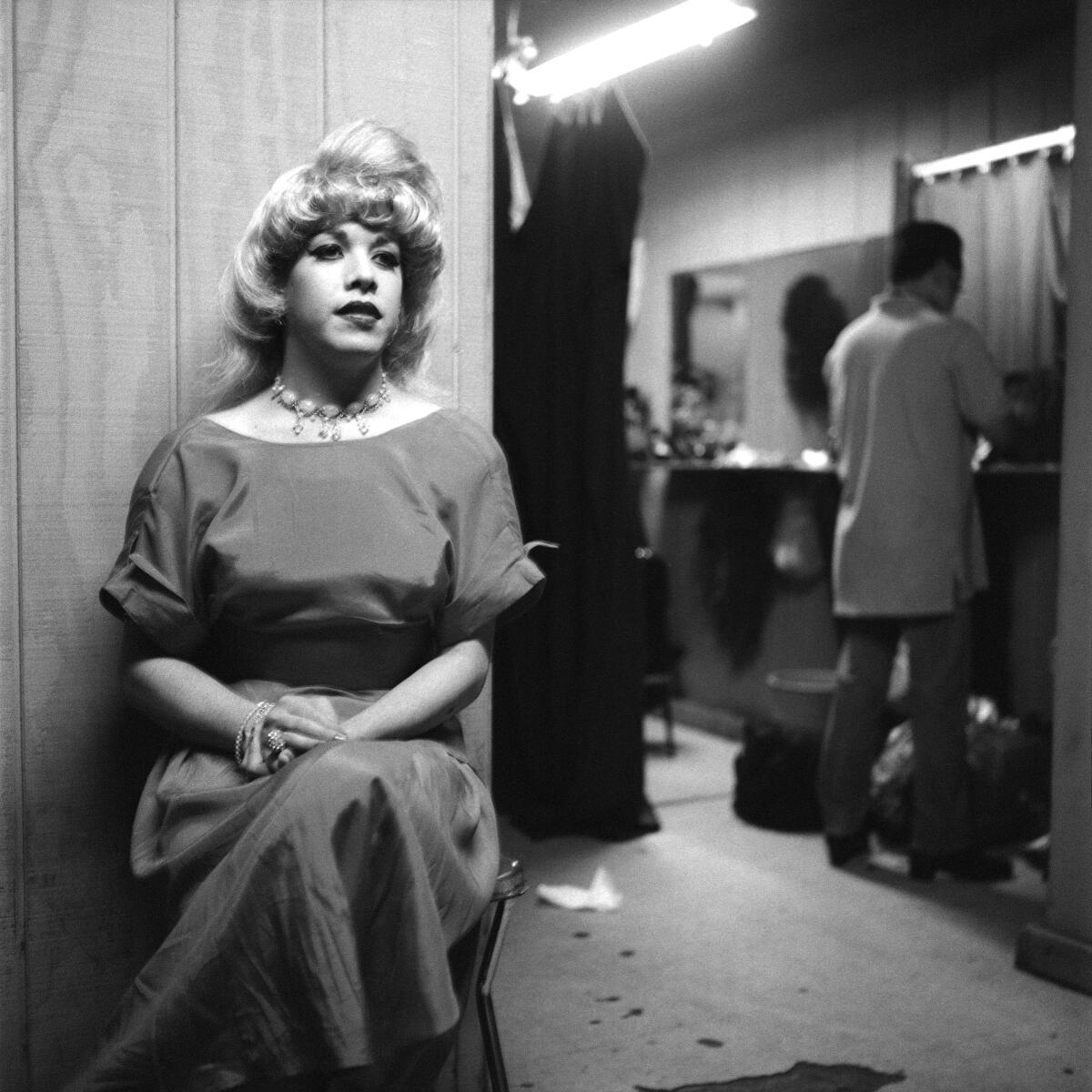 A trans woman in a dress and a blonde wig sits demurely outside of a dressing room