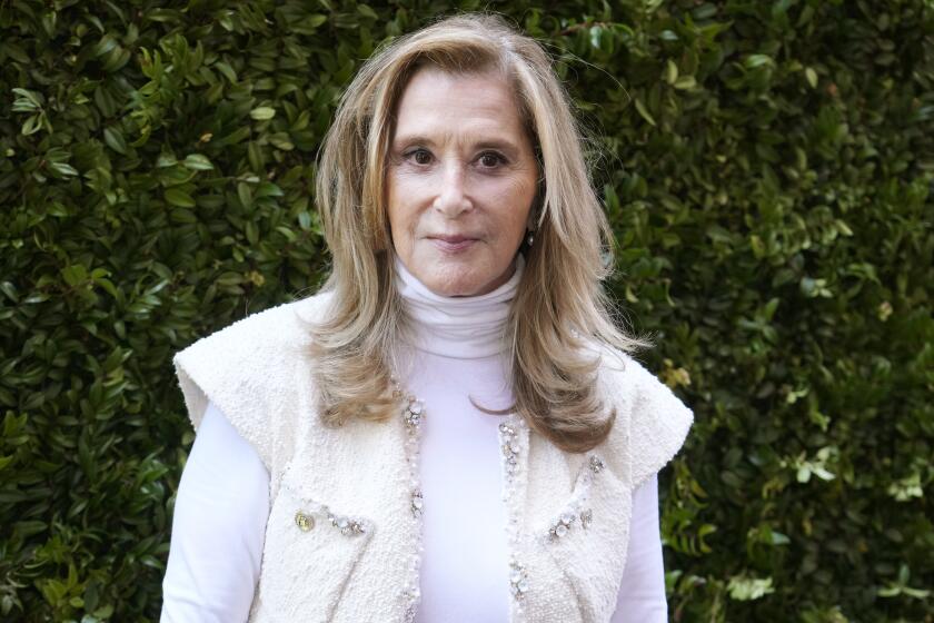 Paula Weinstein wearing a white turtleneck and white vest against some green shrubbery.