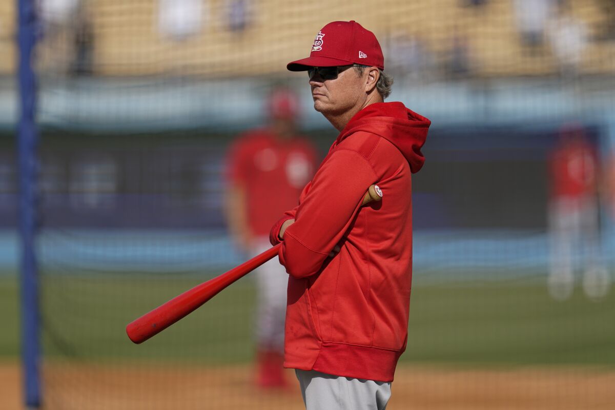 Mike Shildt holds a bat with his arms folded as he stands on the field during batting practice.