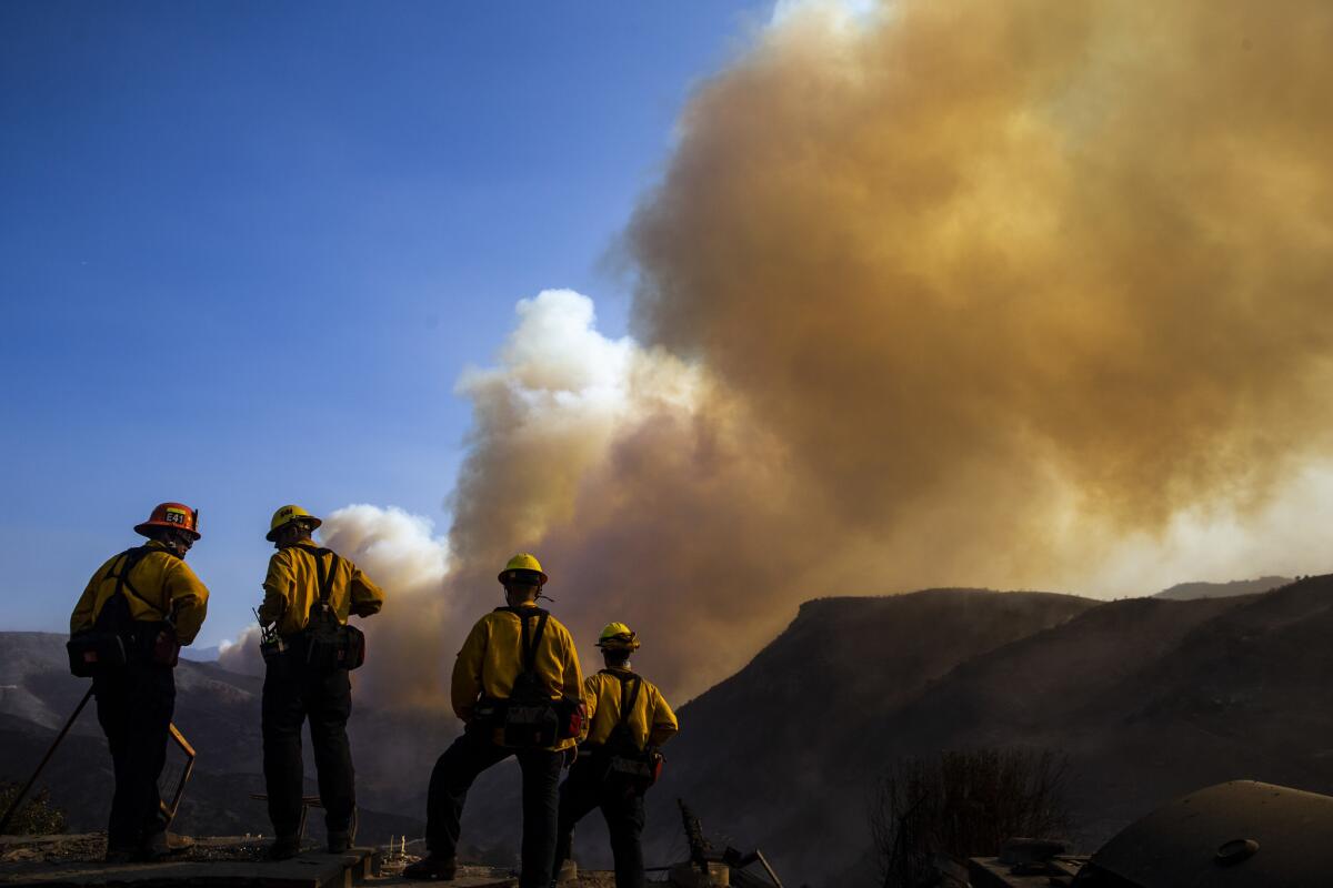 Firefighters working to put out hotspots in and around structures destroyed by the Woolsey fire watch as a plume of smoke rises from near the Chatsworth reservoir.